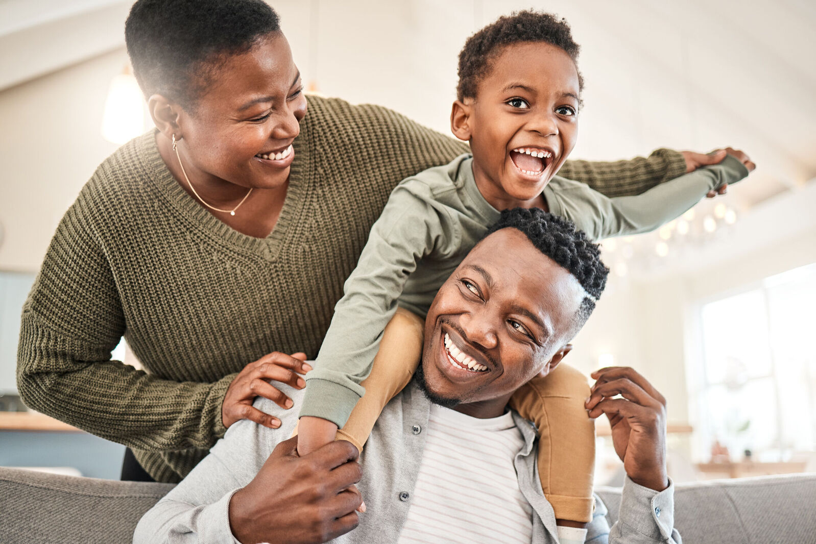 A family relaxes on a couch, with a child playfully perched on one parent's shoulders.
